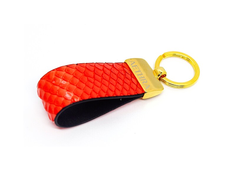 Puede incluir: Llavero de cuero rojo con un anillo dorado y la palabra "AETHIRA" grabada en la etiqueta de metal.