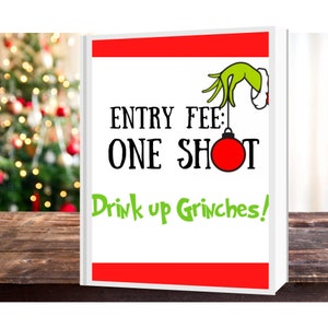 May include: A white sign with red and green accents. The sign says "Entry Fee: One Shot" with a green Grinch hand holding a red ornament. The text at the bottom says "Drink up Grinches!"