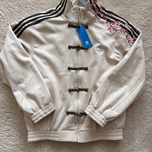Puede incluir: Chaqueta deportiva Adidas color crema con rayas negras en las mangas y un diseño floral en el hombro. La chaqueta tiene cuello mandarín, cierre de cremallera y cierres de palanca. Una etiqueta Adidas está adjunta.