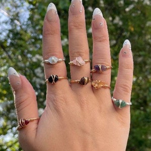 May include: A hand displays a collection of gemstone rings. The rings feature various colored stones, including clear, pink, purple, and green, set in wire. The rings are displayed against a blurred green background.