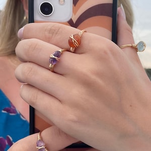 Puede incluir: Primer plano de varios anillos dorados envueltos en alambre con varias piedras preciosas. Los anillos presentan piedras de amatista, cornalina y aguamarina. Los anillos están en una mano, con una funda de teléfono en el fondo.