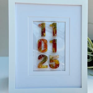May include: A framed artwork featuring the numbers "11 01 25" in a colorful, textured style. The numbers are in shades of red, orange, and yellow, set against a white background within a white frame. The artwork has a modern, decorative aesthetic.