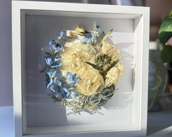 May include: A white frame with a glass front containing a pressed flower arrangement. The arrangement features white and blue flowers, with the white flowers being the most prominent. The flowers are arranged in a circular pattern.