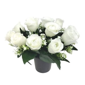 May include: A floral arrangement of white roses, green foliage, and small white flowers. The roses are in full bloom and arranged in a black pot. The arrangement is set against a white background.