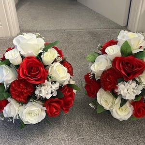 May include: Two circular floral arrangements. Each features red and white roses, red carnations, and white accent flowers. Green leaves are interspersed. The bouquets are placed on a grey surface.