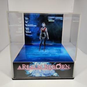 K&ouml;nnte beinhalten: Eine transparente Acryl-Vitrine mit einem Final Fantasy XIV-Diorama. Das Diorama zeigt eine weibliche Figur in roter und schwarzer Kleidung, die auf einer blau beleuchteten Plattform steht. Der Sockel des Geh&auml;uses zeigt das Logo des Spiels.