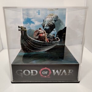 May include: A clear acrylic display case with a 3D pop-up scene of a boat with two figures and a large, gray, mythical creature. The base of the display case has the words "God of War" printed on it.