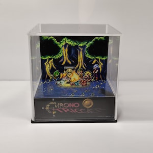 Peut inclure: Diorama en acrylique transparent représentant une scène pixélisée de personnages autour d'un feu de camp dans une forêt. La base affiche le texte "Chrono Trigger" et un motif circulaire.