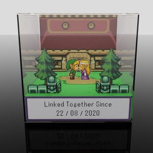 Peut inclure: Un présentoir en acrylique transparent avec une scène pixélisée de deux personnages s'embrassant, avec un bâtiment et des arbres en arrière-plan. Le texte "Linked Together Since 22/08/2020" est affiché sur une étiquette blanche.