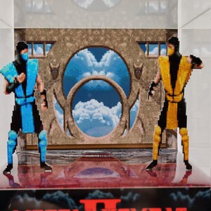 Op de afbeelding: Een display met twee gepixelde figuren uit Mortal Kombat II. E&eacute;n figuur is blauw, de andere geel. De achtergrond toont een gepixelde lucht en een stenen structuur. Het Mortal Kombat II-logo staat onderaan.