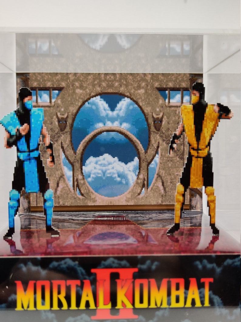 Op de afbeelding: Een display met twee gepixelde figuren uit Mortal Kombat II. E&eacute;n figuur is blauw, de andere geel. De achtergrond toont een gepixelde lucht en een stenen structuur. Het Mortal Kombat II-logo staat onderaan.