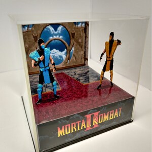 Op de afbeelding: Een Mortal Kombat II-diorama met gepixelde figuren van Sub-Zero en Scorpion in een vitrine. De sc&egrave;ne omvat een rode en grijze vloer, een achtergrond met een lucht en portaal, en het logo van de game.