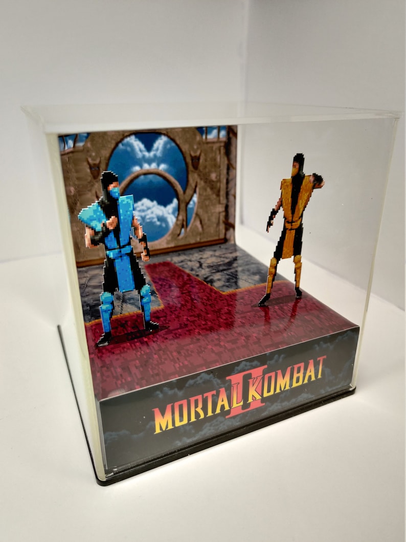 Op de afbeelding: Een Mortal Kombat II-diorama met gepixelde figuren van Sub-Zero en Scorpion in een vitrine. De sc&egrave;ne omvat een rode en grijze vloer, een achtergrond met een lucht en portaal, en het logo van de game.