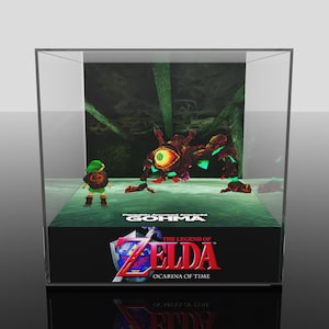 Puede incluir: Una vitrina que presenta una escena de The Legend of Zelda: Ocarina of Time. La escena incluye al personaje Link, un gran enemigo Gohma y el logotipo del juego. La vitrina es transparente, con una base negra y una superficie reflectante.