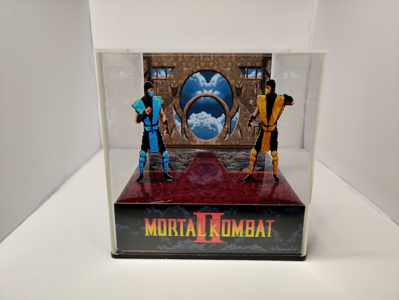 Op de afbeelding: Een diorama met twee gepixelde figuren uit Mortal Kombat II, &eacute;&eacute;n in blauw en de andere in geel, tegen een achtergrond van een portaal en lucht. De basis toont het logo van de game.