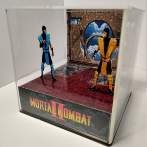 Op de afbeelding: Een Mortal Kombat II diorama met gepixelde figuren van Sub-Zero en Scorpion in een doorzichtige vitrine. De basis toont het logo van het spel en een rode vloer. De achtergrond toont een portaal met uitzicht op de hemel.