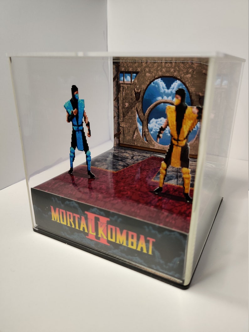 Op de afbeelding: Een Mortal Kombat II diorama met gepixelde figuren van Sub-Zero en Scorpion in een doorzichtige vitrine. De basis toont het logo van het spel en een rode vloer. De achtergrond toont een portaal met uitzicht op de hemel.