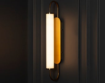 Puede incluir: Una lámpara de pared moderna con acabado dorado y una fuente de luz cilíndrica vertical. La lámpara tiene una parte superior arqueada y curvada y una placa trasera rectangular. La luz emite un brillo cálido.