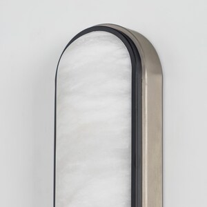 May include: A vertical wall-mounted light fixture with an arched top. The fixture features a central panel of white, marbled glass encased in a thin black frame, which is surrounded by a wider, brushed metallic silver outer border.