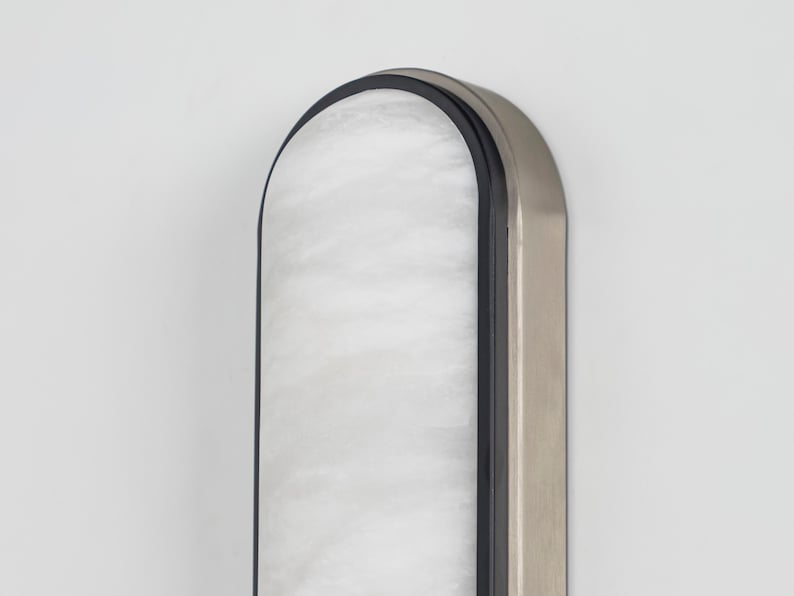 May include: A vertical wall-mounted light fixture with an arched top. The fixture features a central panel of white, marbled glass encased in a thin black frame, which is surrounded by a wider, brushed metallic silver outer border.