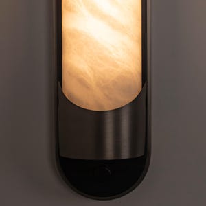 May include: A vertical wall sconce featuring a cylindrical, translucent, cream-colored shade with a marbled texture. The shade is partially encased in a dark, matte-finished metal frame with a curved, semi-circular lower support and a circular button.