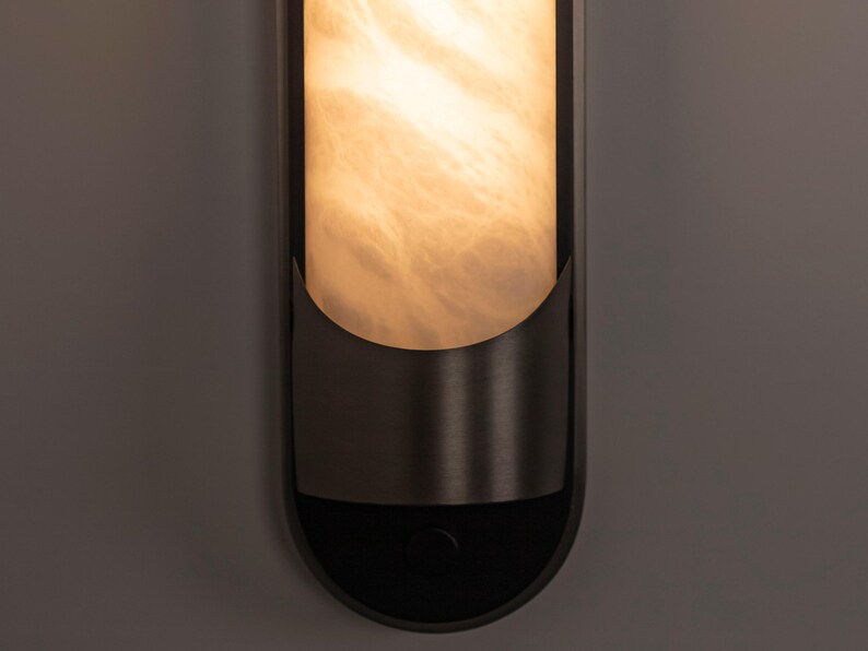 May include: A vertical wall sconce featuring a cylindrical, translucent, cream-colored shade with a marbled texture. The shade is partially encased in a dark, matte-finished metal frame with a curved, semi-circular lower support and a circular button.