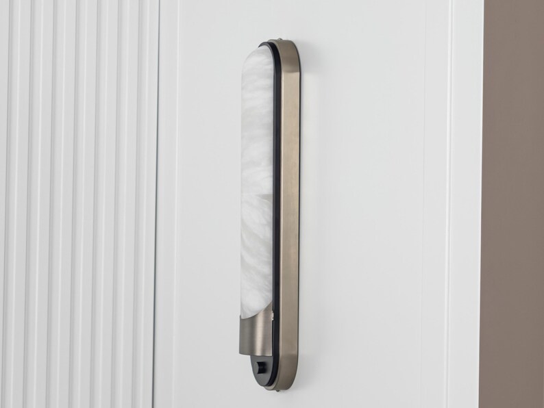 May include: A vertical wall-mounted light fixture featuring a long, rounded rectangular frame in a metallic bronze finish. The center panel consists of a translucent, white, marbled glass diffuser that emits a soft, diffused light glow.