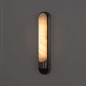 May include: A vertical wall sconce featuring a long, rounded rectangular frame in a dark metallic finish. The center panel is made of a translucent, marbled white material that emits a warm, soft glow, creating a diffused light effect.