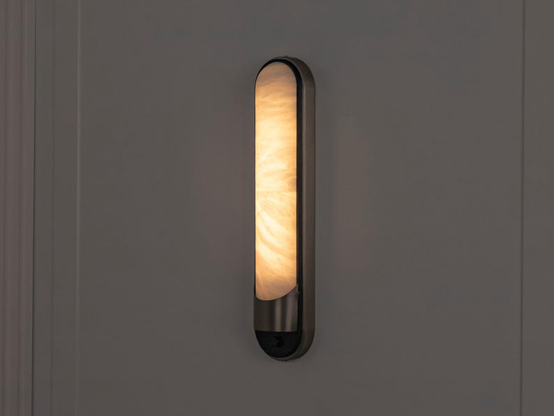 May include: A vertical wall sconce featuring a long, rounded rectangular frame in a dark metallic finish. The center panel is made of a translucent, marbled white material that emits a warm, soft glow, creating a diffused light effect.