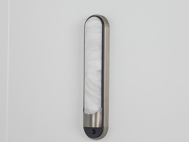 May include: A vertical wall sconce featuring a brushed nickel frame and a long, slender, white marbled glass diffuser. The fixture has a rounded top and a bottom section with a small black circular button, showcasing a clean, elongated design.
