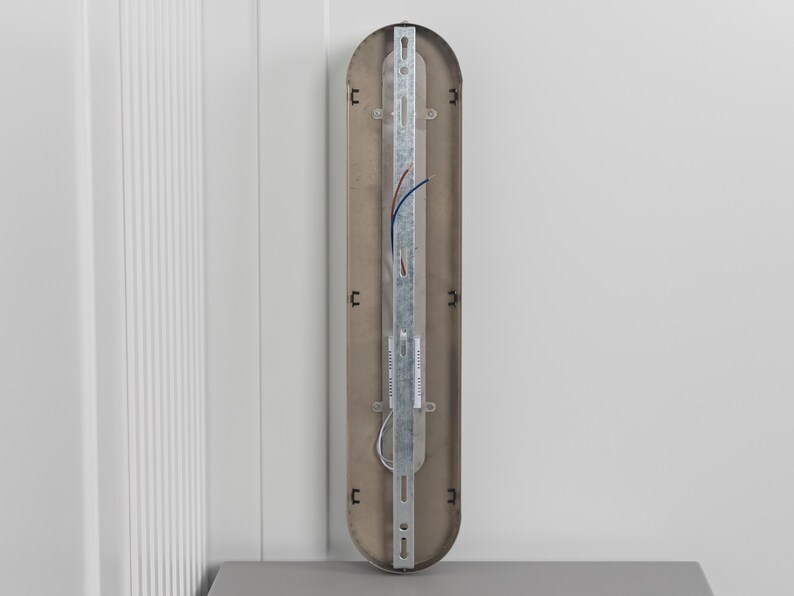 May include: A vertical, pill-shaped light fixture backplate with a metallic finish. It features a central galvanized steel mounting bracket, exposed electrical wiring in blue and red, and small black plastic clips along the inner edges for assembly.