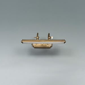 May include: A brass-toned picture light fixture with a rectangular base and a horizontal light bar. The fixture has two adjustable arms and a white light source. The design is classic and elegant, suitable for illuminating artwork or decor.