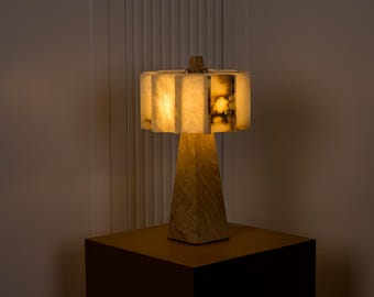 May include: A decorative table lamp with a geometric base and a segmented, illuminated shade. The shade is made of a translucent material, possibly alabaster or onyx, and emits a warm, inviting glow. The lamp is placed on a dark brown surface.