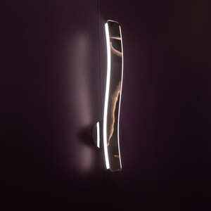 Puede incluir: Una moderna lámpara de pared curvada con un acabado de metal pulido y una brillante luz LED lineal. La lámpara está montada en una pared de color púrpura oscuro, creando un contraste con la luz.