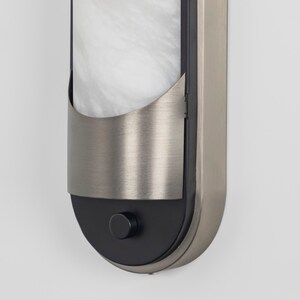 May include: A vertical wall sconce featuring a brushed nickel frame with a rounded bottom. The fixture includes a white marble-patterned glass panel partially covered by a curved metal shield and a small black circular knob at the base.