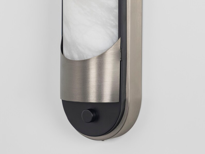 May include: A vertical wall sconce featuring a brushed nickel frame with a rounded bottom. The fixture includes a white marble-patterned glass panel partially covered by a curved metal shield and a small black circular knob at the base.