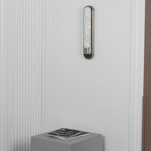 May include: A vertical wall-mounted light fixture with a brushed metal frame and a long, rounded, translucent white glass diffuser. The fixture features a small black circular sensor at the bottom and emits a soft, diffused glow.