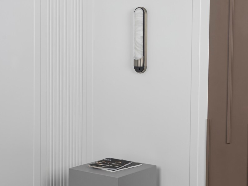 May include: A vertical wall-mounted light fixture with a brushed metal frame and a long, rounded, translucent white glass diffuser. The fixture features a small black circular sensor at the bottom and emits a soft, diffused glow.