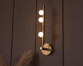 May include: A modern wall sconce with a brass finish. The fixture features three spherical light bulbs arranged vertically along a slender rod. The sconce is mounted on a circular base and is set against a brown wall.
