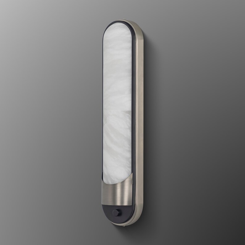 May include: A vertical wall sconce featuring a long, rounded rectangular shape with a white, marbled glass diffuser. The fixture has a brushed metallic frame and a dark gray base section at the bottom, which includes a small, circular push-button switch.