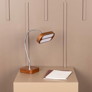 May include: A wooden desk lamp with a flexible, silver-colored neck and a rectangular base. The lamp head has two white LED lights. A notebook rests on the table next to the lamp.