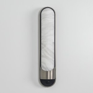 May include: A vertical wall-mounted light fixture with a long, rounded rectangular shape. It features a white, marbled glass diffuser panel framed in dark metal, with a brushed silver metal accent at the base and a small circular black button.