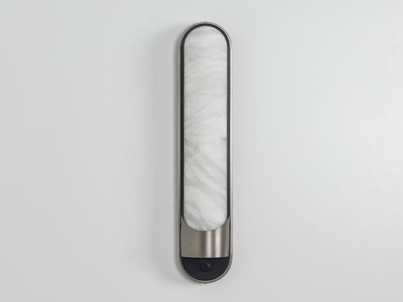 May include: A vertical wall-mounted light fixture with a long, rounded rectangular shape. It features a white, marbled glass diffuser panel framed in dark metal, with a brushed silver metal accent at the base and a small circular black button.