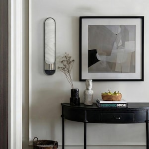 May include: A black semi-circular console table holds a woven basket of green pears, a stack of books including one titled Mykonos Muse, and a stone sculpture. A wall-mounted light fixture and a framed abstract painting hang above the table.