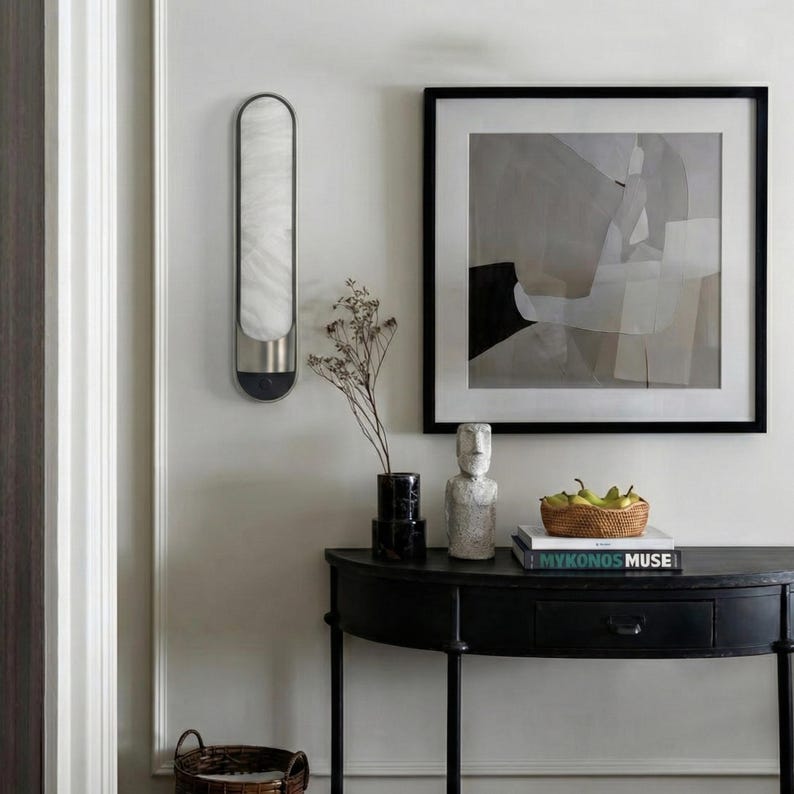 May include: A black semi-circular console table holds a woven basket of green pears, a stack of books including one titled Mykonos Muse, and a stone sculpture. A wall-mounted light fixture and a framed abstract painting hang above the table.