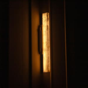 Könnte beinhalten: Eine rechteckige Wandleuchte mit warmem, leuchtendem Licht. Die Leuchte hat einen langen, vertikalen, strukturierten, cremefarbenen Schirm und eine dunkle Metallhalterung. Das Licht ist an einer dunkelbraunen Wand montiert.
