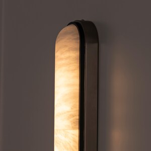 May include: A vertical wall sconce featuring a long, arched, translucent panel with a marbled, cream-colored texture that emits a warm, soft glow. The light fixture is encased in a dark, metallic frame with rounded edges and a smooth finish.