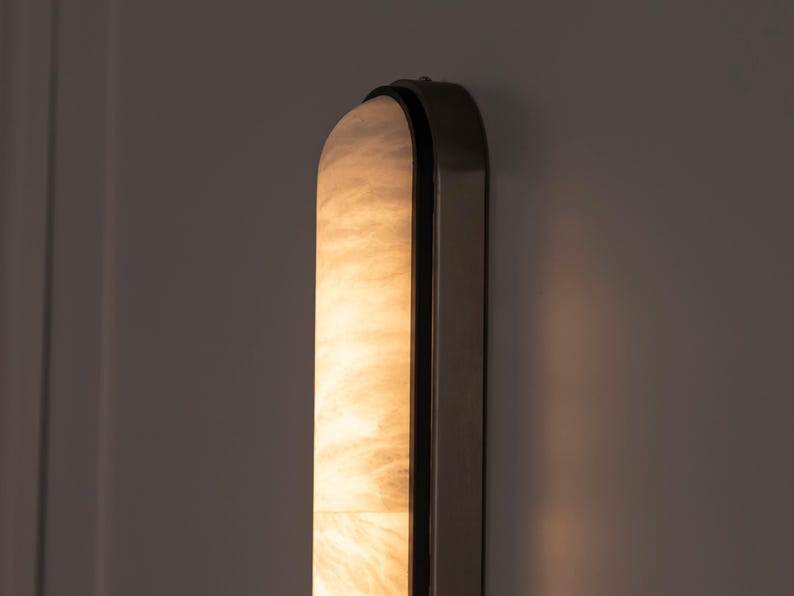 May include: A vertical wall sconce featuring a long, arched, translucent panel with a marbled, cream-colored texture that emits a warm, soft glow. The light fixture is encased in a dark, metallic frame with rounded edges and a smooth finish.