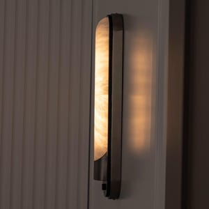 May include: A vertical wall sconce featuring a long, rectangular, translucent shade with a marbled, cream-colored pattern. The fixture is encased in a dark metal frame with a smooth finish, emitting a warm, soft glow from the illuminated panel.