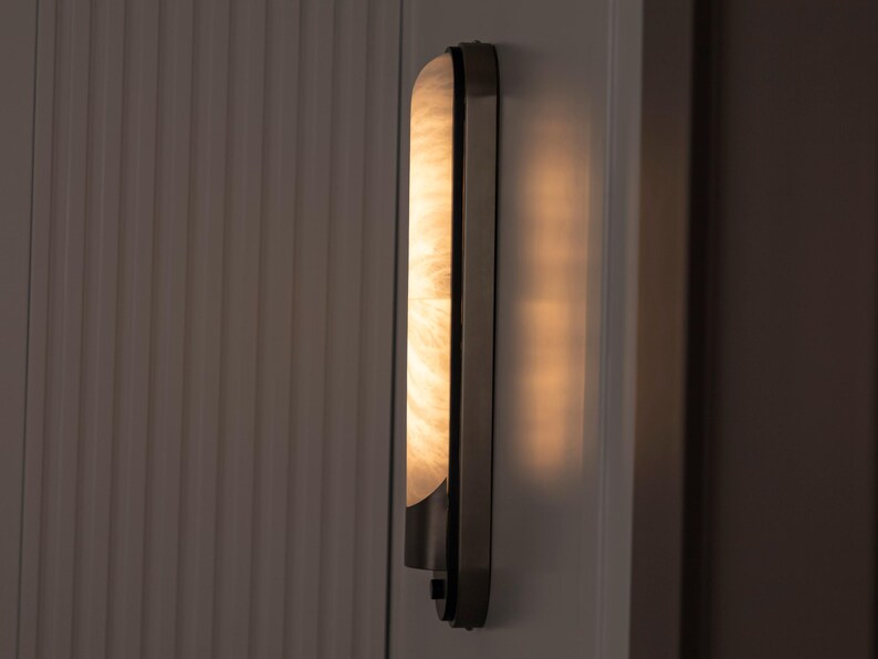 May include: A vertical wall sconce featuring a long, rectangular, translucent shade with a marbled, cream-colored pattern. The fixture is encased in a dark metal frame with a smooth finish, emitting a warm, soft glow from the illuminated panel.